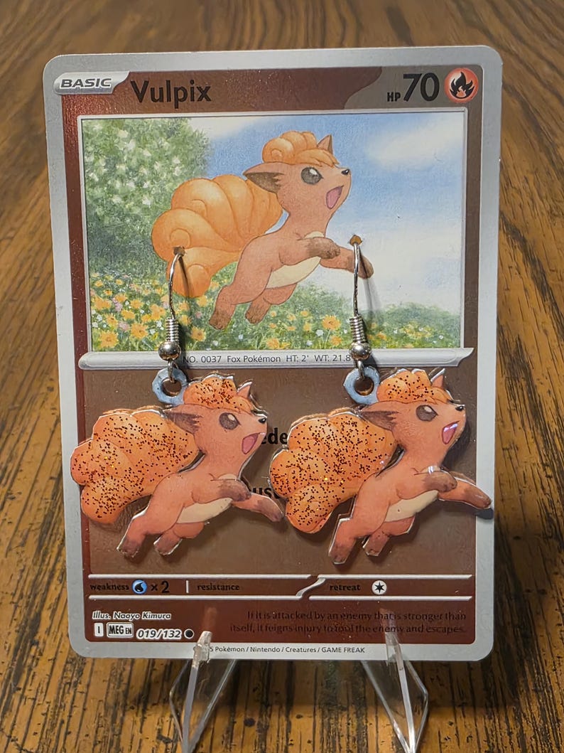 May include: A pair of dangle earrings featuring Vulpix, a tan and orange fox-like creature with a multi-tailed design and glitter accents. The earrings are displayed against a Vulpix trading card with the text Vulpix, 70 HP, and Basic.