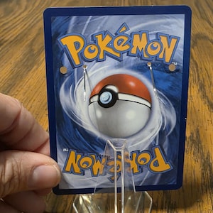 May include: A rectangular display card featuring the blue and yellow Pok&eacute;mon logo at the top and bottom, with a central red and white Pok&eacute; Ball graphic against a swirling blue background. Two silver metal earring hooks are attached to the card.