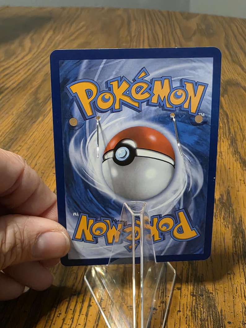 May include: A rectangular display card featuring the blue and yellow Pok&eacute;mon logo at the top and bottom, with a central red and white Pok&eacute; Ball graphic against a swirling blue background. Two silver metal earring hooks are attached to the card.