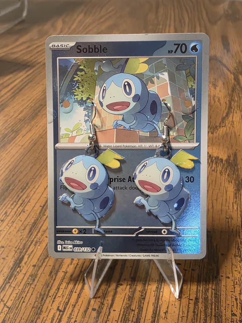 Peut inclure: Une paire de boucles d'oreilles pendantes repr&eacute;sentant le personnage bleu et bleu clair Sobble de la franchise Pok&eacute;mon. Les boucles d'oreilles sont pr&eacute;sent&eacute;es sur une carte &agrave; collectionner Sobble, affichant le personnage, le nom Sobble et le texte HP 70.