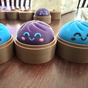 May include: A collection of rounded, dome-shaped figures with smiling faces, each seated in a brown cylindrical base. The figures are colored in vibrant shades of blue and purple, featuring simple, hand-painted facial expressions with eyes and cheeks.