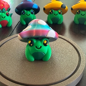 May include: A collection of small, green frog figurines, each wearing a different colored mushroom cap. The foreground features a frog with a multi-colored, gradient mushroom cap with white spots, while others in the back display red, purple, and yellow caps.