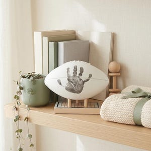 May include: A white football featuring a dark gray handprint on its side, mounted on a light-colored wooden stand with a grid base. The football is displayed on a shelf alongside a green potted plant, books, and a folded cream-colored knit blanket.