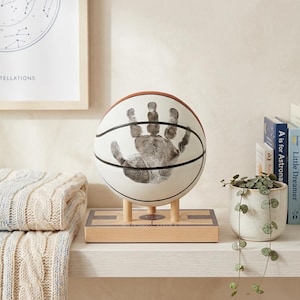 May include: A white basketball featuring a black handprint graphic, mounted on a light-colored wooden stand designed like a basketball court. The stand includes a small circular logo and text that reads STAY SMALL DREAM BIG.