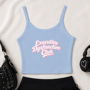 Puede incluir: Un top corto sin mangas de color azul claro con tirantes finos. El centro de la prenda presenta el texto "Executive Dysfunction Club" impreso en una fuente blanca estilizada con un contorno rosa suave, creando un diseño gráfico claro.