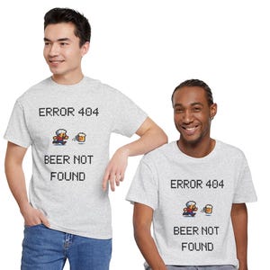 Könnte beinhalten: Ein hellgraues Kurzarm-T-Shirt mit einer Pixel-Art-Grafik, die eine Person zeigt, die auf einen Bierkrug zuläuft. Der Text ERROR 404 BEER NOT FOUND ist in einer schwarzen, verpixelten Schriftart mittig auf der Brust des Kleidungsstücks platziert.
