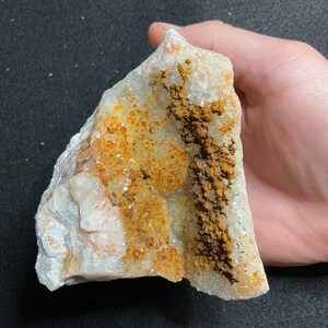 May include: A raw, translucent white quartz crystal specimen featuring a prominent, textured vertical band of dark brown iron oxide staining and patches of vibrant orange mineral deposits across its crystalline, jagged surface structure.