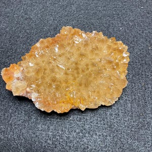 May include: A cluster of golden-orange citrine crystals with a jagged, irregular shape. The surface is composed of numerous small, pointed, translucent crystal points that catch the light, creating a sparkling, multifaceted appearance.
