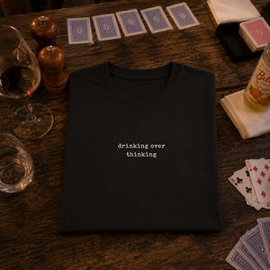 May include: A black crew neck t-shirt featuring the white text "drinking over thinking" printed in a typewriter-style font centered on the chest. The shirt is folded neatly, showcasing a simple design with short sleeves and a standard collar.