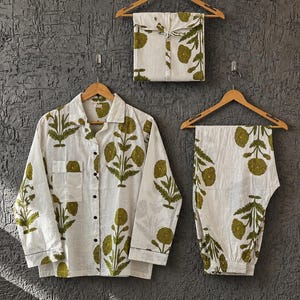 Könnte beinhalten: Ein Set aus einem weißen, langärmeligen Hemd mit Knopfleiste und passender Hose, verziert mit einem olivgrünen floralen Blockdruckmuster. Das Set enthält ein gefaltetes Kleidungsstück, alles auf Holzkleiderbügeln vor einer dunklen Wand.