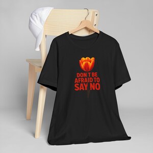 May include: A black short-sleeved t-shirt featuring a graphic of an orange and red tulip flower above the red text that reads, "DON'T BE AFRAID TO SAY NO." The shirt is displayed on a wooden hanger, and a white baseball cap is draped over the chair.