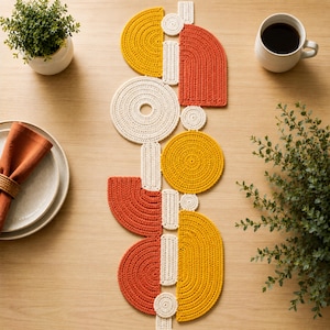 Può includere: Un runner da tavola lungo e stretto realizzato con forme geometriche all'uncinetto nei colori giallo senape, arancione bruciato e crema. Il design presenta cerchi, semicerchi e connettori rettangolari interconnessi in un motivo verticale.
