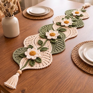 Può includere: Un lungo runner da tavola all'uncinetto smerlato con un motivo ripetuto di sezioni in pizzo color crema e foglie verde scuro. Fiori bianchi tridimensionali con centri gialli sono applicati lungo il centro, rifiniti con nappe color crema.