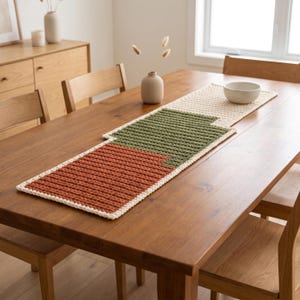 Puede incluir: Un camino de mesa rectangular de ganchillo con un diseño de bloques de color en hilo crema, verde oliva y naranja quemado. El camino presenta un patrón de punto acanalado y texturizado, extendido sobre el centro de una mesa de comedor de madera.