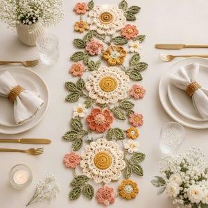 Könnte beinhalten: Ein gehäkelter Tischläufer mit einem Blumenmuster aus großen cremefarbenen Blüten, kleineren rosa, gelben und orangefarbenen Blumen sowie grünen Blättern. Der Läufer liegt auf einem Tisch mit weißen Tellern, goldenem Besteck und Gläsern.