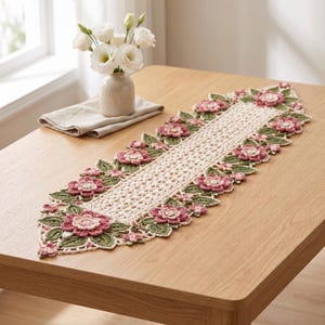 Puede incluir: Un camino de mesa de ganchillo de color crema, de forma alargada, con un patrón central calado bordeado por flores rosas en relieve y hojas verdes. Los motivos florales están dispuestos simétricamente a lo largo de los bordes.