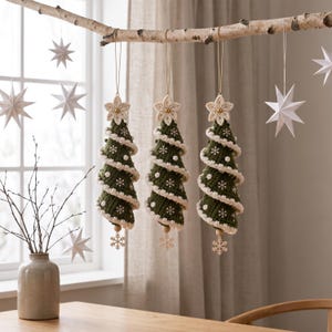May include: Three hanging ornaments shaped like spiral Christmas trees made of dark green crocheted yarn with white trim, small white pom-poms, and embroidered snowflake patterns. Each tree is topped with a cream crocheted star and a wooden bead.