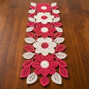 May include: A rectangular crochet table runner featuring a repeating floral pattern. The design consists of alternating large white and dark pink flowers with contrasting centers, surrounded by smaller crocheted leaves in matching colors.