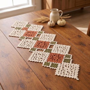 May include: A crochet table runner featuring a geometric pattern of cream, burnt orange, and olive green square motifs. The runner is composed of intricate floral-inspired lace squares joined together to create a long, decorative centrepiece.