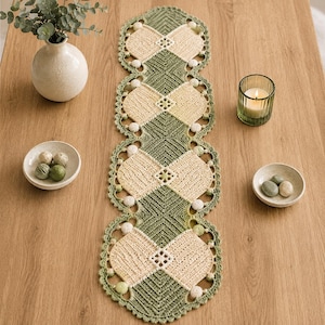 May include: A long, rectangular crochet table runner featuring a repeating geometric pattern in sage green and cream yarn. The runner has scalloped edges decorated with small, round, cream-colored pom-poms and a central openwork diamond design.