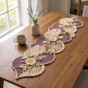Puede incluir: Un camino de mesa de ganchillo largo y decorativo con un patrón intrincado de motivos circulares alternos en color crema y morado polvoriento. El camino está adornado con flores de ganchillo tridimensionales en crema y hojas verde salvia.