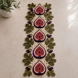 May include: A rectangular crochet table runner featuring a central column of five stylized fig halves in shades of deep purple, pink, and gold. The runner is bordered by intricate cream-colored lace and alternating green fig leaves and small purple buds.