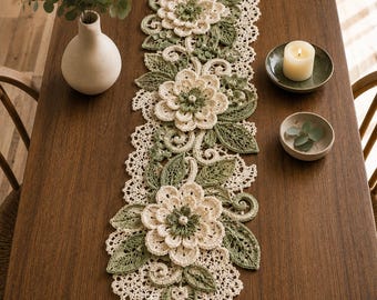 May include: A long, rectangular crochet table runner featuring a cream and sage green floral pattern. The design includes three large, layered crochet flowers surrounded by intricate green leaves, swirling vines, and a delicate lace-like border.