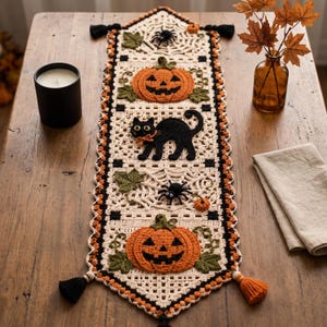 May include: A rectangular crochet table runner featuring a cream-colored base with black and orange accents. The design includes two orange jack-o'-lanterns, a black cat, two black spiders, green leaves, and web patterns, finished with tassels.
