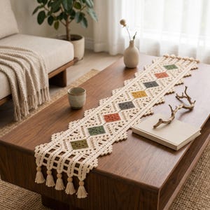 May include: A long, cream-colored crochet table runner featuring a geometric pattern of diamond-shaped motifs in olive green, terracotta, and mustard yellow. The runner has a scalloped edge, intricate openwork, and ends with wooden beads and tassels.