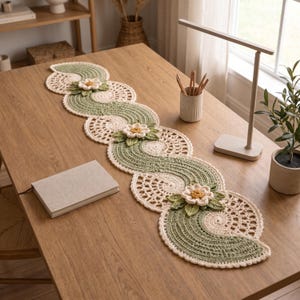 Può includere: Un lungo runner da tavola all'uncinetto a forma di serpente con un motivo ripetuto di fasce ondulate verde salvia e motivi circolari in pizzo color crema. Fiori tridimensionali color crema con centri gialli e foglie verdi sono applicati.