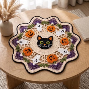 May include: A round, crocheted doily featuring a central black cat face with green eyes, surrounded by alternating orange jack-o'-lanterns and white ghosts. The outer edge is decorated with purple spider web patterns and green leaf accents.