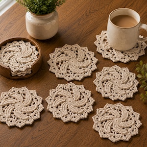 May include: A set of round, beige crochet coasters featuring a spiral pattern and scalloped edges. The coasters are arranged individually and in a stack within a circular wooden holder, with one coaster placed under a ceramic mug filled with coffee.