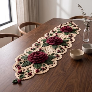 Puede incluir: Un camino de mesa de ganchillo color crema con tres rosas grandes y tridimensionales de color rojo oscuro y varios capullos pequeños. El camino tiene un borde festoneado y está decorado con hojas de ganchillo verde oscuro en el encaje.