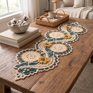 May include: A long, crocheted table runner featuring a repeating pattern of cream-colored circular doilies, dark teal leaves, and mustard yellow flowers. The intricate lace design is laid flat across a rustic wooden table surface.