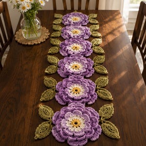 May include: A long crochet table runner featuring a repeating pattern of six large, layered purple flowers with white and yellow centers. Each flower is flanked by two textured green leaves, creating a continuous floral design along the length.