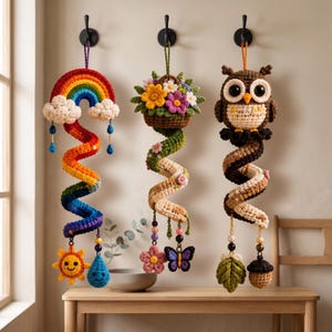May include: Three hanging crochet decorations featuring a rainbow with clouds and a sun, a basket of colorful flowers with a butterfly, and a brown owl with an acorn and leaf. Each piece has a spiral body and hangs from a black wall hook.