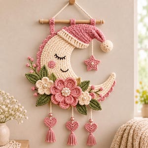 Pode incluir: Uma tapeçaria de crochê com uma lua crescente cor de creme com rosto adormecido e gorro rosa. Decorada com flores rosas e creme, folhas verdes e três corações pendentes com borlas e contas, suspensa por uma haste de madeira.