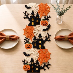 May include: A Halloween-themed crochet table runner featuring black haunted houses, orange spider webs, white circular doilies, orange jack-o'-lanterns, and black bats. The runner is arranged vertically with two plates and orange napkins on the sides.