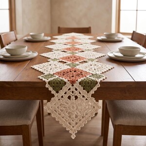 May include: A rectangular crochet table runner featuring a repeating pattern of square motifs in cream, sage green, and terracotta orange. The runner has an intricate, openwork lace design with scalloped edges, laid flat along the centre of a table.