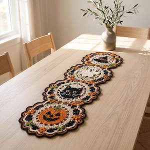 Peut inclure: Un chemin de table en crochet composé de quatre motifs circulaires reliés entre eux. Les motifs représentent une citrouille d'Halloween, une tête de chat noir, un fantôme blanc et un chapeau de sorcière, réalisés en fil orange, noir, blanc et vert.