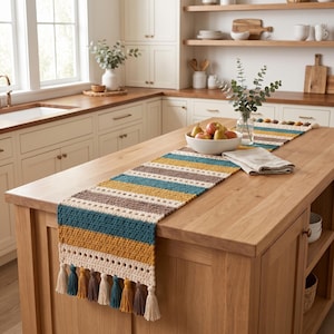 Puede incluir: Un camino de mesa rectangular de ganchillo con rayas horizontales en azul cerceta, amarillo mostaza, marrón y crema. El camino presenta un patrón de punto texturizado y está rematado con borlas alternas en crema, azul cerceta, marrón y amarillo mostaza.