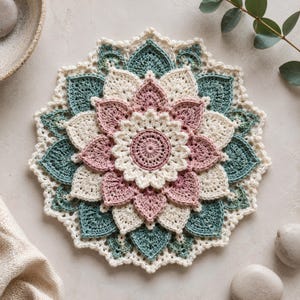 May include: A circular crochet mandala doily featuring a layered floral design. The center is dusty rose, surrounded by alternating petals in cream and dusty rose, with an outer border of teal and cream leaf-shaped motifs in a textured stitch.