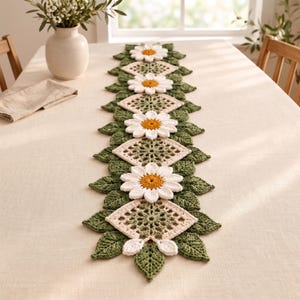 Può includere: Un runner da tavola rettangolare all'uncinetto con un motivo ripetuto di margherite bianche con centri gialli, alternate a pannelli quadrati in pizzo color crema, il tutto bordato da foglie all'uncinetto testurizzate verde scuro.
