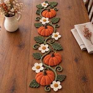May include: A long, crocheted table runner featuring a repeating pattern of orange pumpkins, white flowers with yellow centres, and dark green leaves connected by winding vines. The textile decor is laid out vertically on a wooden surface.