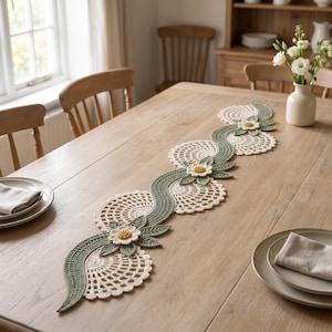 Op de afbeelding: Een lange, decoratieve gehaakte tafelloper met een ingewikkeld, kronkelend rankpatroon in saliegroen, geaccentueerd met crèmekleurige cirkelvormige kantmotieven en driedimensionale witte bloemen met gele harten, plat op een houten tafel.