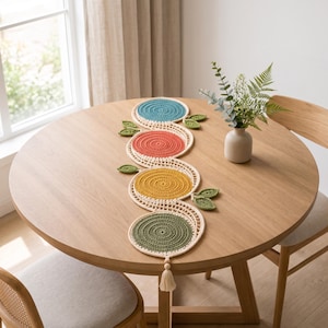 May include: A crochet table runner featuring four circular motifs in teal, coral, mustard yellow, and olive green, connected by a cream-colored lattice border. The runner is accented with small green crocheted leaves and a single cream tassel at one end.