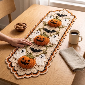 Op de afbeelding: Een rechthoekige gehaakte tafelloper met een crèmekleurige basis en een geschulpte rand in oranje en zwart. De loper is versierd met driedimensionale oranje pompoenen, witte spookapplicaties en zwarte vleermuismotieven verbonden door groene ranken.
