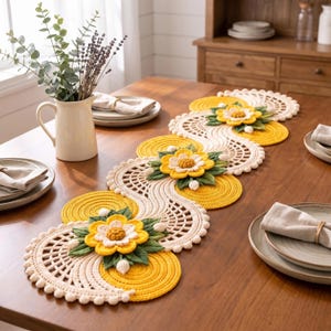 May include: A long, serpentine table runner featuring a crochet design with alternating cream and yellow circular patterns. The runner is decorated with three-dimensional yellow flowers with white centers and green leaves, finished with small cream pom-poms.