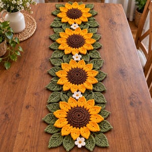 May include: A crochet table runner featuring four large sunflowers with dark brown centers and bright yellow petals, surrounded by green crocheted leaves and small white flowers arranged in a linear pattern along the center of the table.
