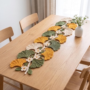 Può includere: Un runner da tavola all'uncinetto con un motivo ripetuto di foglie di ginkgo nei toni del giallo senape, verde salvia e crema, arricchito da piccoli fiori bianchi. Il runner è disposto lungo un tavolo da pranzo in legno chiaro.